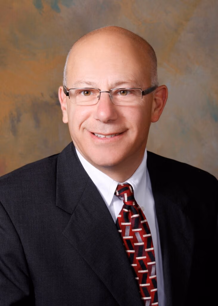 Portrait of a smiling, bald man wearing glasses, a dark suit, and a red patterned tie against a neutral backdrop.