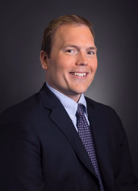Professional headshot: smiling man in a navy suit and purple patterned tie against a dark gradient background.