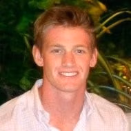 Smiling man with short blond hair wearing a light shirt, outdoors with greenery behind him.