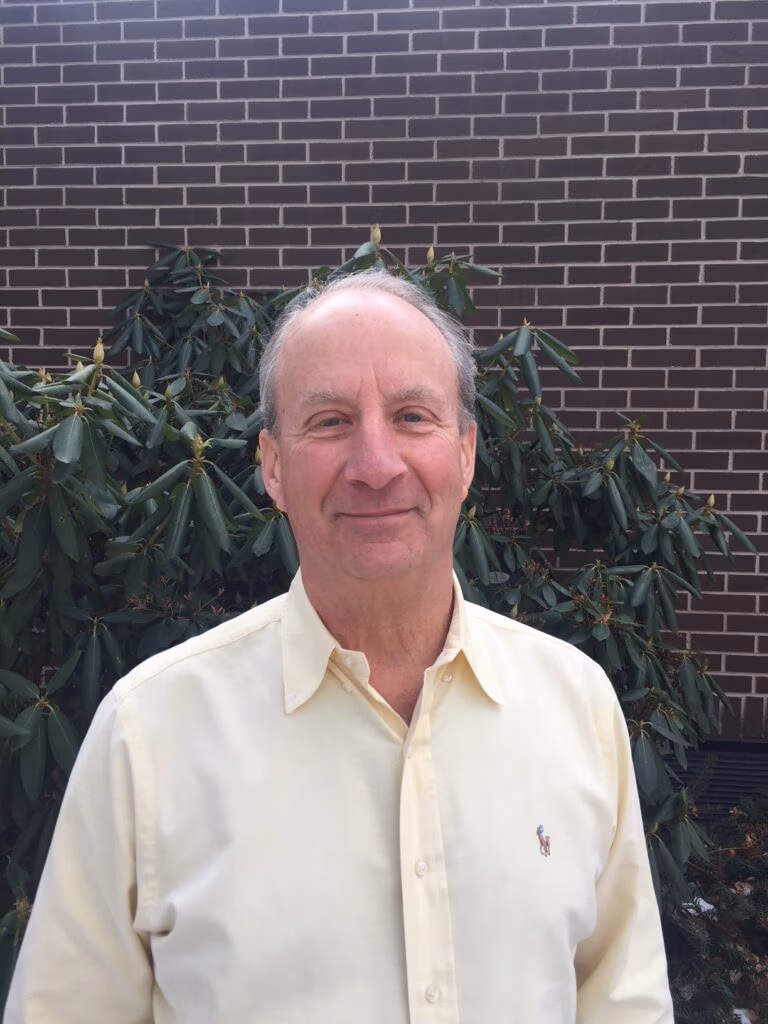 Smiling man in a pale yellow shirt standing in front of a brick wall with green shrubs behind him.