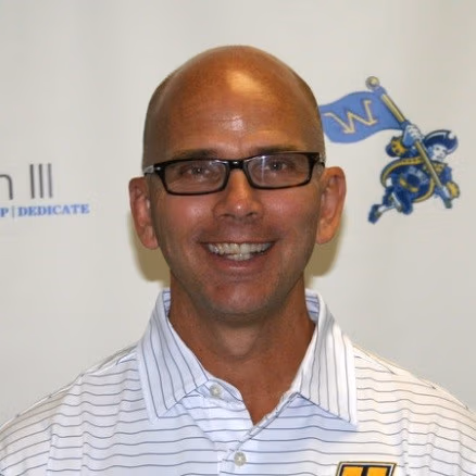 Smiling bald man with glasses in a striped shirt, head-and-shoulders portrait against a light background with logos behind him.