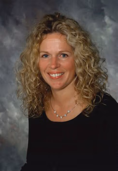 Portrait of a smiling woman with curly blonde hair, wearing a black top and a beaded necklace, posed against a gray studio backdrop.