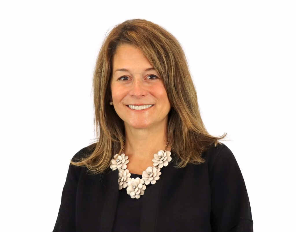 Smiling woman with shoulder-length light brown hair wearing a black blazer and a white flower necklace.