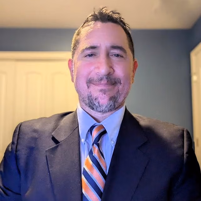 Smiling man in a dark suit and striped orange, blue, and white tie, posing in a blue-walled room with white cabinets in the background.
