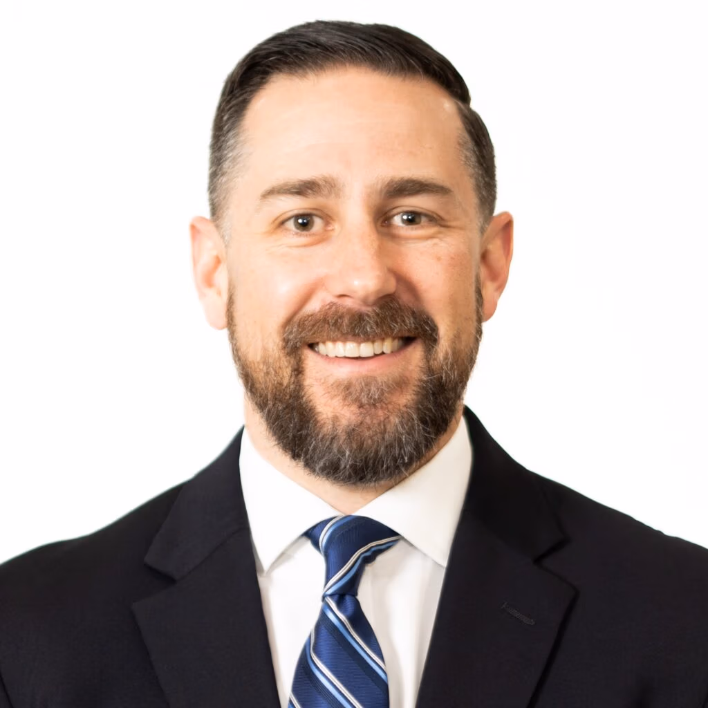 Partial portrait of a man in a dark suit and blue striped tie, cropped at the shoulders, smiling