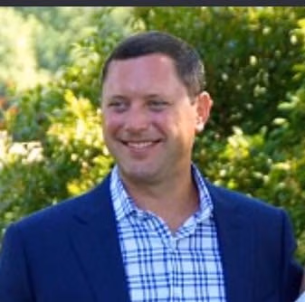 Smiling man in a blue blazer and checkered shirt outdoors with greenery in the background.