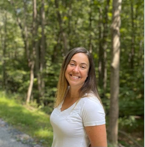 Smiling woman with light brown hair wearing a white shirt standing on a forest path surrounded by trees.