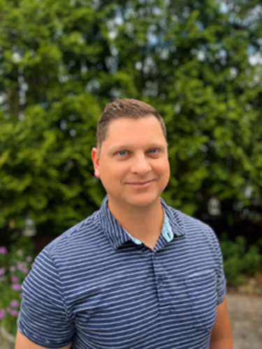 Smiling man in a blue striped polo shirt outdoors with green bushes behind him.
