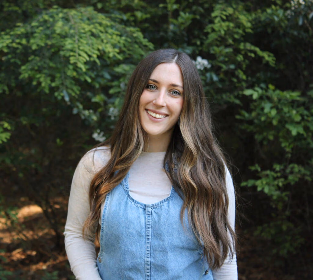 Smiling young woman with long brown hair outdoors, wearing a light gray top under a denim jumper, green foliage behind.