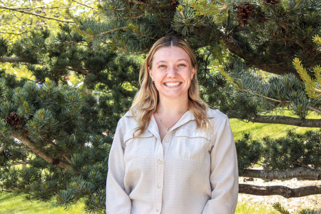 Smiling woman standing outdoors with pine tree branches behind her and sunlight.