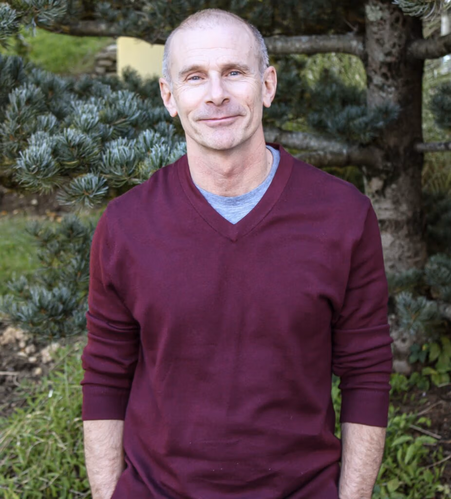 Middle-aged man in a maroon sweater standing outdoors with green shrubs behind him.