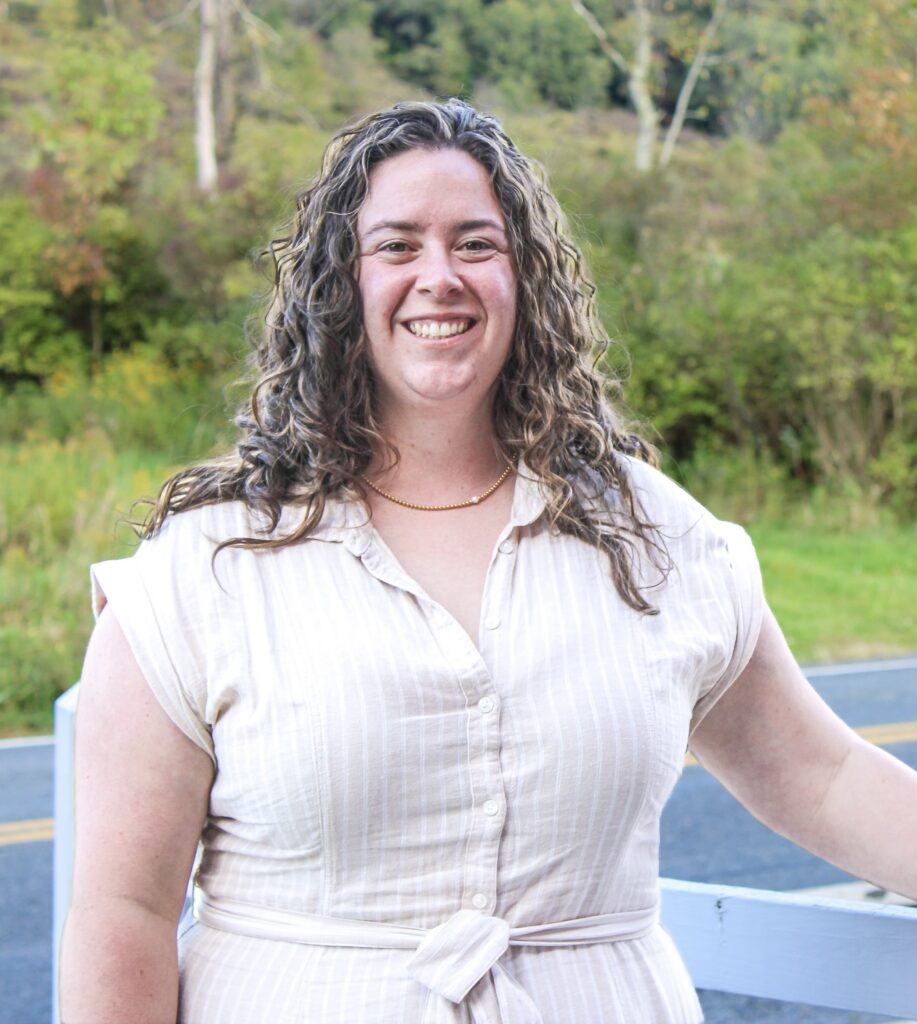 Smiling woman with curly hair wearing a light striped dress outdoors, greenery in the background.